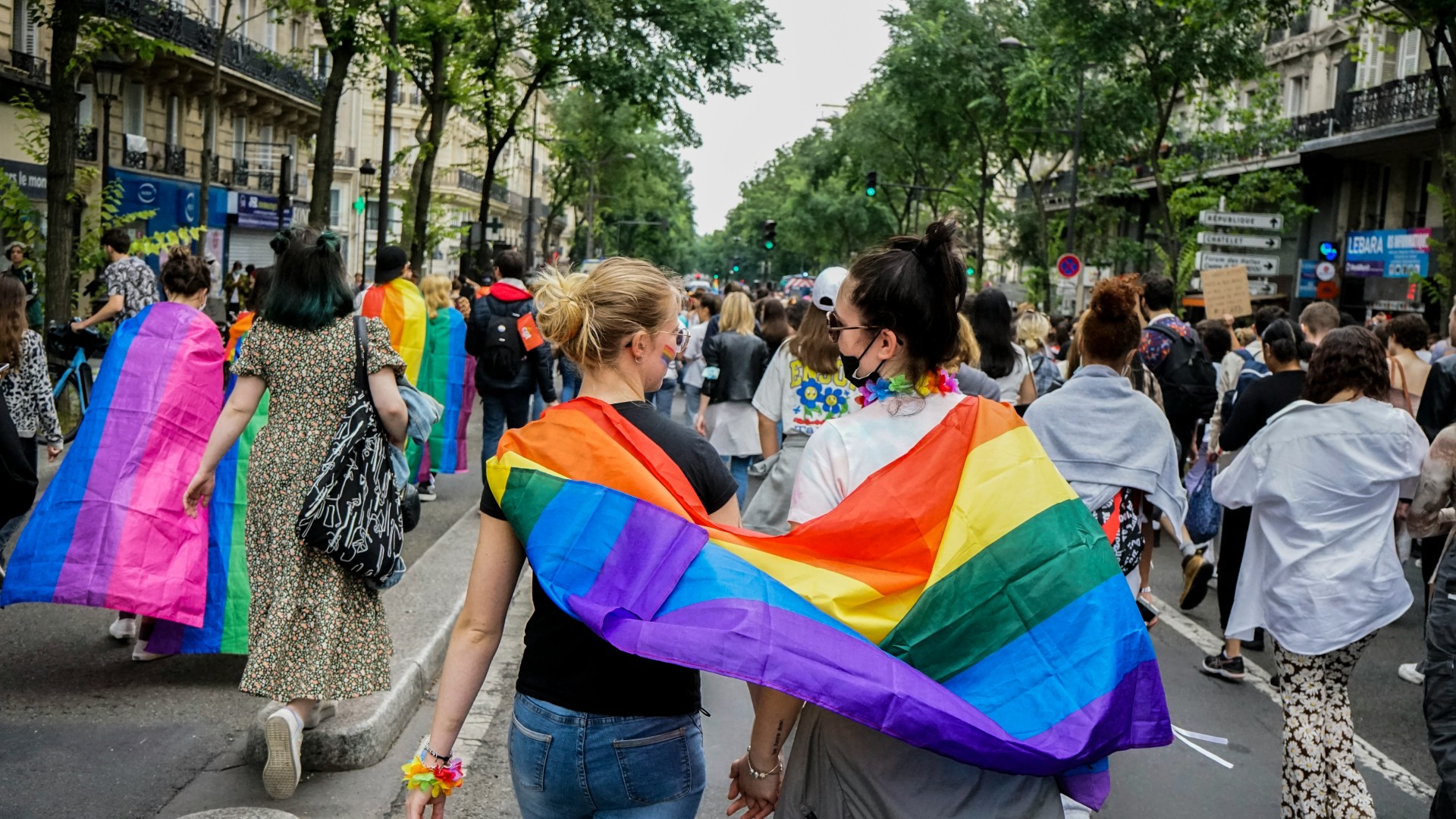 Due persone sotto una bandiera LGBTQIA+ a un corteo durante Pride Month.
