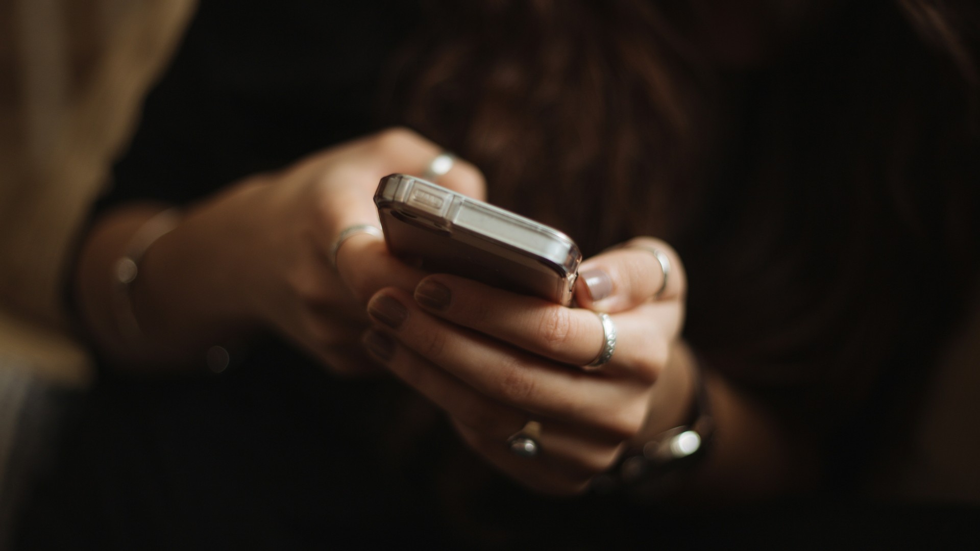 A phone in the hands of a young woman who wears a black top in the background.
