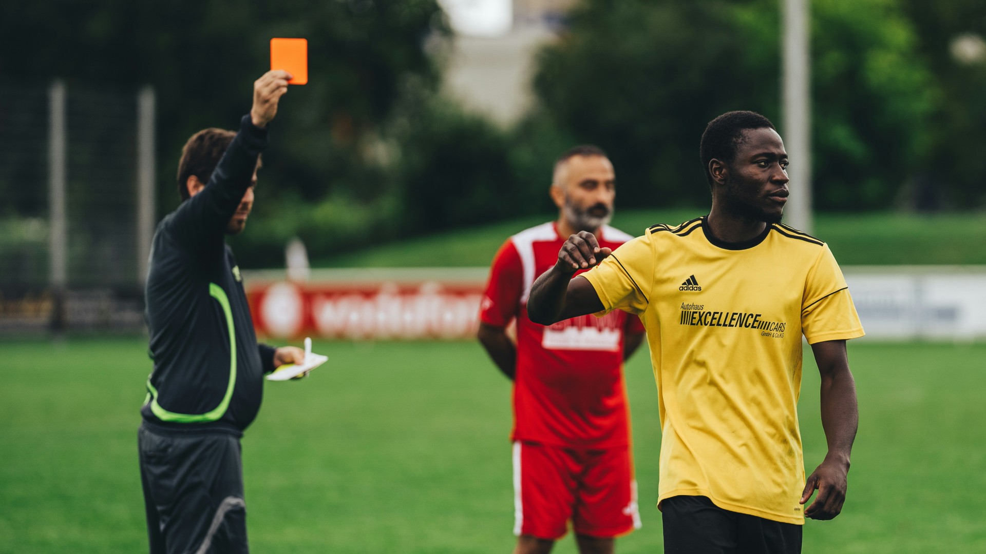 Two football players and a referee holding an orange card against one of them.